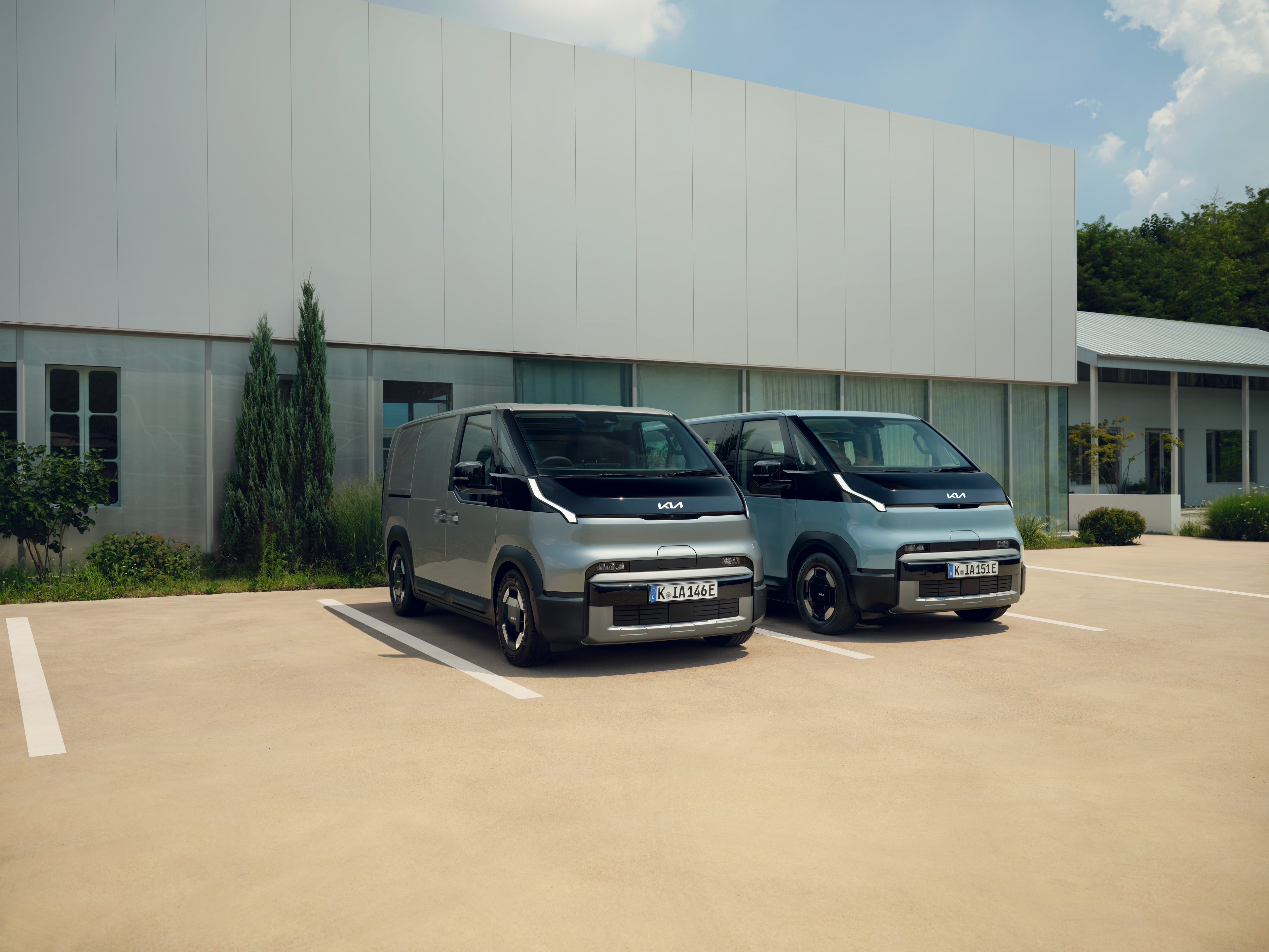 Two Kia PV5 vans, a grey Cargo and a light blue Passenger, parked side by side outside a modern building